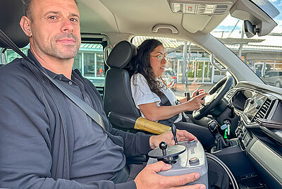 Zum Fenster der Beifahrerseite sieht man zwei Personen in einem Auto sitzen. Auf der Beifahrerseite sitzt einer unserer Fahrlehrer der PARAVAN Fahrschule und auf dem Fahrersitz, sitzt eine Fahrschülerin, die gerade trotz neuromuskulären Erkrankungen lernt, mit PARAVAN Fahrhilfen Auto zu fahren.