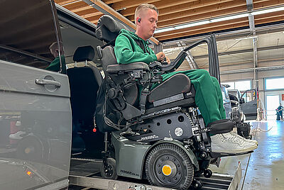 Mann mit Tetraplegie fährt im Elektrorollstuhl über einen Fahrzeuglift in ein barrierefrei umgebautes Auto.