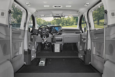 Der hintere Innenraum eines VW New Caravelle, bei dem man zur einen Seitentüre reinschaut und durch die andere geöffnete Türe den herausgefahrenen Kassettenlift mit Rollstuhl davor sieht. Mann kann ebenfalls im Innenraum des Autos sehen das es kein Beifahrersitz gibt, sowie das der Boden Tiefergelegt worden ist im Vergleich zur Heck Bank.