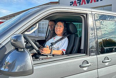 Sabina sitzt auf dem Fahrersitz eines umgebauten Fahrzeugs vor einem Gebäude mit dem Schriftzug „PARAVAN“; das Fahrzeug ist für barrierefreies Fahren angepasst.