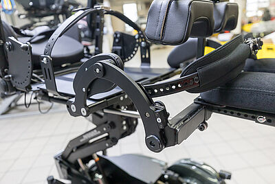 Detailaufnahme eines komplexen mechanischen Gelenks und Sitzsystems eines Elektrorollstuhls, mit gepolsterten Armlehnen und Kopfstütze.