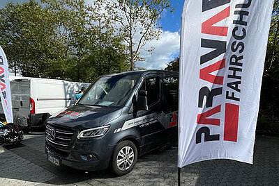 Eine weiße Paravanfahne mit Logo der Fahrschule weht vor dem Fahrschulsprinter