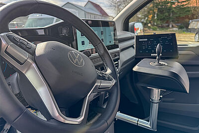 Ein Detailbild des Innenraus des neuen VW New Caravelle, bei dem man klare Sicht auf das Lenkrad hat, von der Fahrerseite aus. Etwas unscharf im Hintergrund sieht man ein eingebauter Paravan Joystick mit PARAVAN Touch Display.