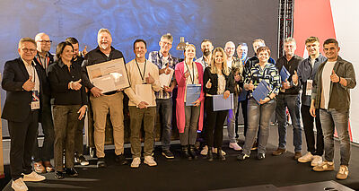 Gruppenfoto von Mitarbeitenden bei einer Firmenveranstaltung. Die Teilnehmenden stehen auf einer Bühne, halten Urkunden und Auszeichnungen und zeigen überwiegend den Daumen nach oben.