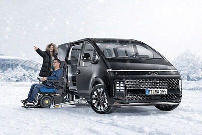 Frontansicht eines umgebauten Paravan Hyundai Staria mit herausgefahrenem Kassettenlift und einem Paar in einer Winterlandschaft. der Mann sitzt dabei im Rollstuhl