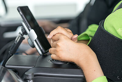 Zwei Hände halten einen Joystick fest mit dem derjenige sein Auto steuert, trotz Muskelerkrankung. direkt daneben befindet sich sein Handy mit der er seine Sekundärfuktionen steuern kann. 