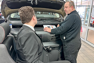 Zwei Männer im Gespräch vor einem Fahrzeug mit geöffneter Motorhaube, einer davon im Rollstuhl mit Anzug, der andere in Arbeitskleidung.