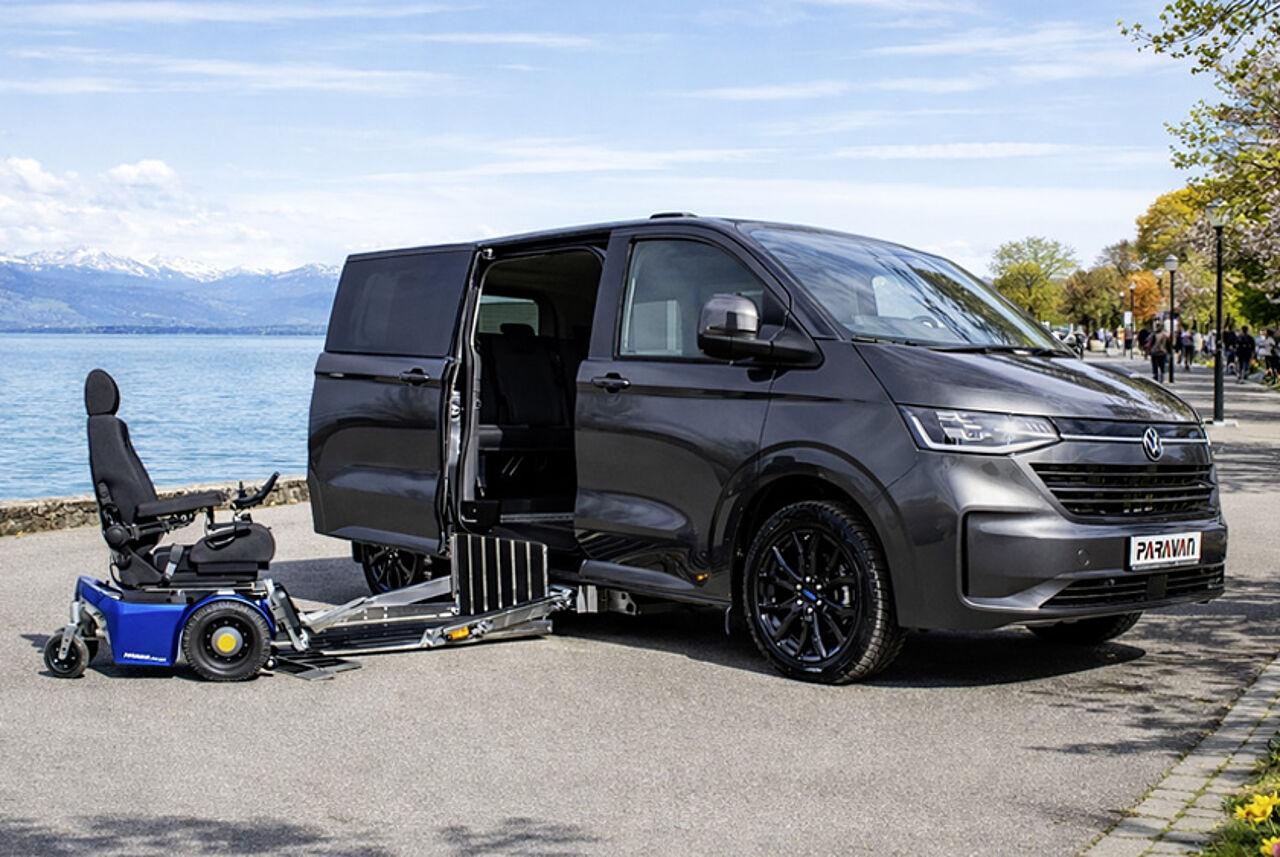 Der PARAVAN VW Caravelle Umbau steht an der Promenade eines Sees, davor ein blauer Elektrorollstuhl