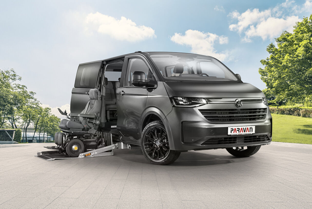 Der PARAVAN VW Caravelle Umbau steht an der Promenade eines Sees, davor ein blauer Elektrorollstuhl