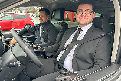 Ein lächelnder junger Mann sitzt in einem Anzug, in seinem Auto. Er macht gerade seine Fahrausbildung in der PARAVAN Fahrschule, denn neben ihm sitzt der Fahrlehrer.Er fährt über zwei Joysticks mit dem Drive-by-Wire System Space Drive von PARAVAN.