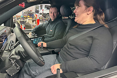 Eine lachende Frau mit Muskelschwäche sitzt auf dem Fahrersitz eines von PARAVAN umgebauten Fahrzeugs, mit Joystick-Steuerung und digitalem Drive-by-Wire System. Auf dem Beifahrersitz, sitzt einer unserer Fahrlehrer.