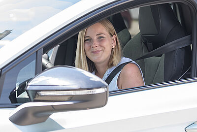 Eine lächelnde junge Frau sitz am Steuer eines weißen PKW und schaut zum offenen Fenster heraus.