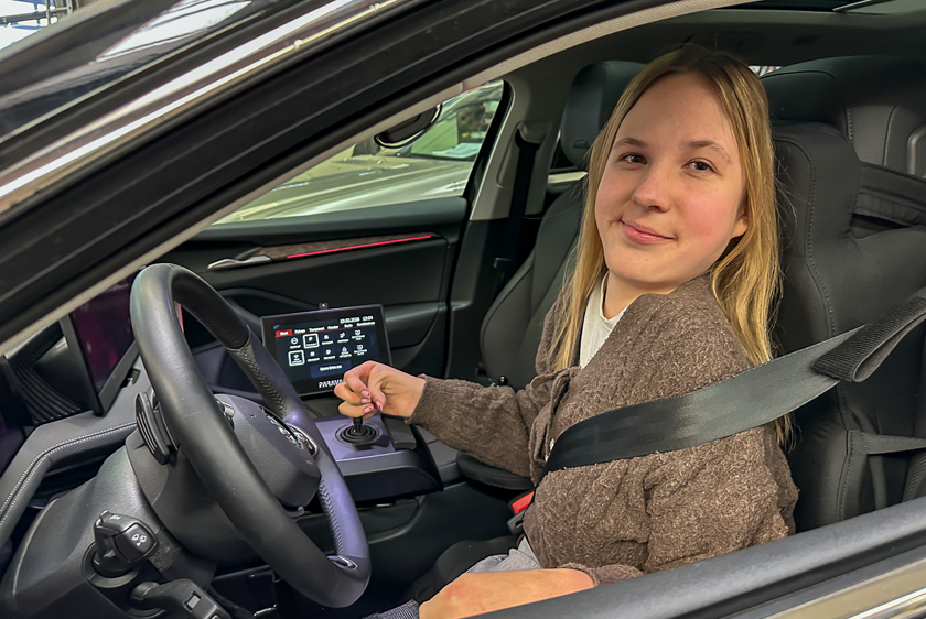 Eine lächelnde junge Dame mit blonden Haaren, sitzt auf dem Fahrersitz eines Autos und schaut zum geöffneten Fenster hinaus. In ihrer rechten Hand hält sie ein Joystick fest. 