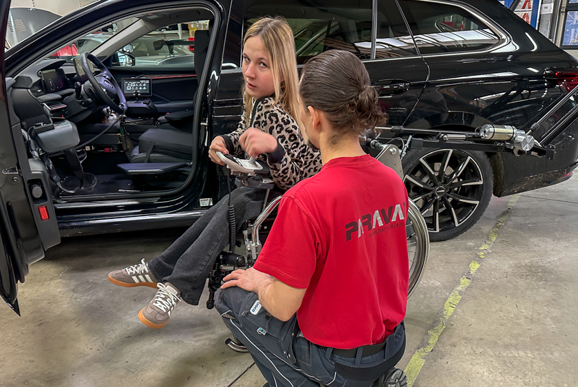 Eine junge blonde Dame sitzt in ihrem Rollstuhl, parallel zu einem schwarzen Fahrzeug, bei dem die Fahrertüre offen ist. Aus der Heckklappe kommt ein Roboterarm, auf der rechten Seite des Bildes, zum Rollstuhl. Vor ihr Kniet ein Mann mit einem roten Paravan T-Shirt. 