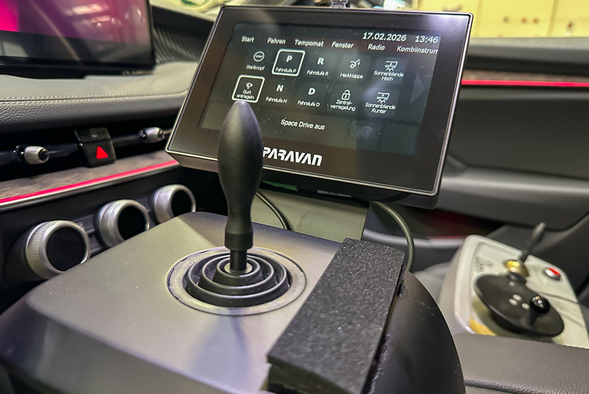Im Vordergrund sieht man einen Joystick verbaut in einem Auto. Hinter dem Joystick sieht man ein Paravan Touch Display, welches verschiedene Funktionen zum Autofahren aufzeigt. 