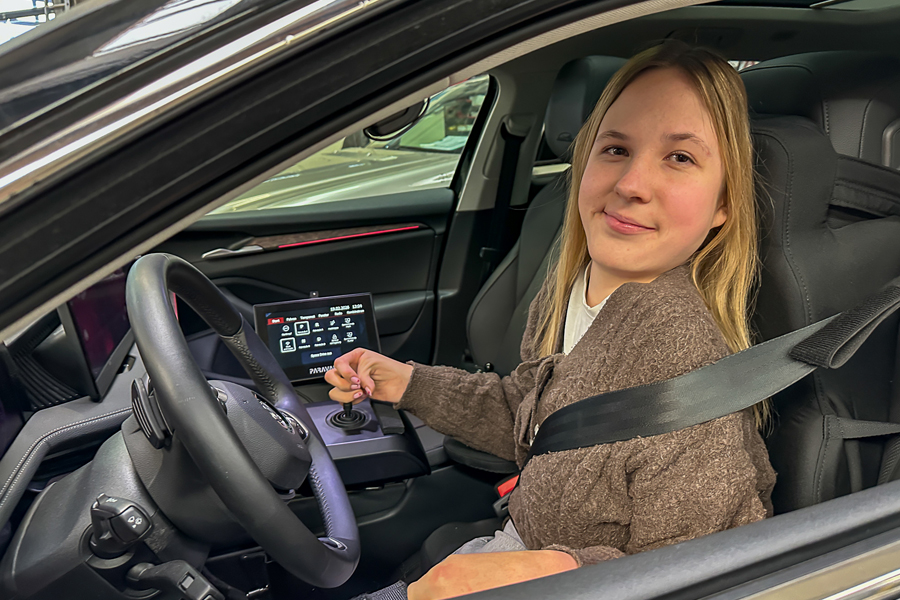 Eine lächelnde junge Dame mit blonden Haaren, sitzt auf dem Fahrersitz eines Autos und schaut zum geöffneten Fenster hinaus. In ihrer rechten Hand hält sie ein Joystick fest. 