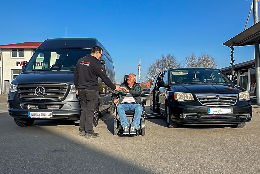 Bei blauem Himmel stehen zwei Fahrzeuge, links ein neuer Sprinter, rechts ein Chrysler Voyager auf dem Hof von Paravan. Zwischen den beiden Fahrzeugen sitzt ein Mann lachend im Rollstuhl, dem gerade ein Mitarbeiter von Paravan die Hand schüttelt.