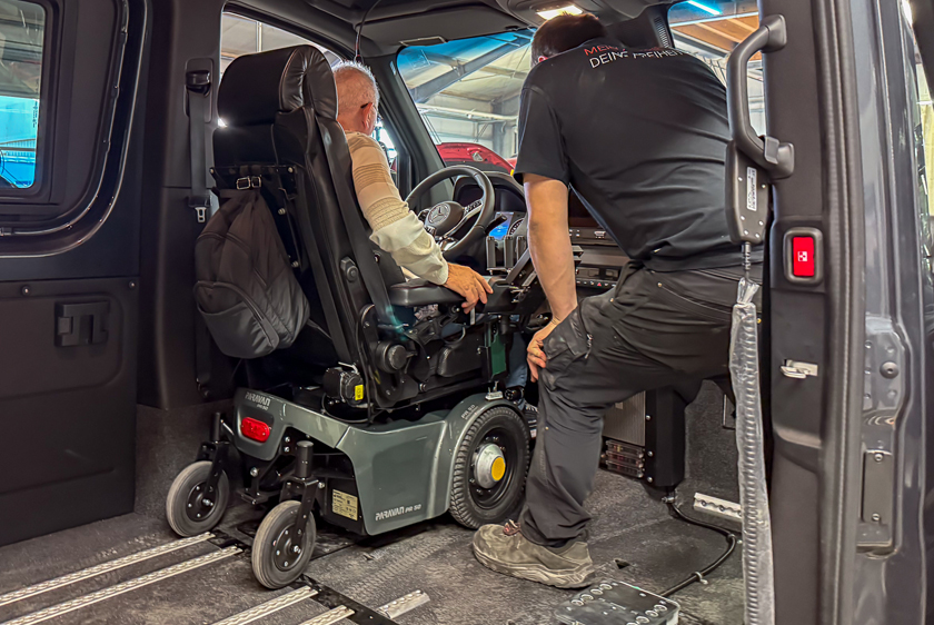 Ein Mann in einem Paravan PR50 Elektrorollstuhl, steht vor dem Lenkrad in einem Sprinter.  Anstelle des Beifahrersitz, steht dort ein Paravan Mitarbeiter, der prüfend schaut.