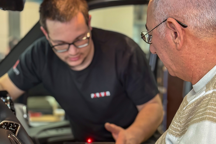 Im Vordergrund befindet sich links ein Lenkrad eines Autos und rechts ein Mann mit weißen kurzen haaren, einer Brille und einem braun gestreiften Pullover. Im Hintergrund, unscharf, erklärt ein Paravan Mitarbeiter das Paravan Touch Display.