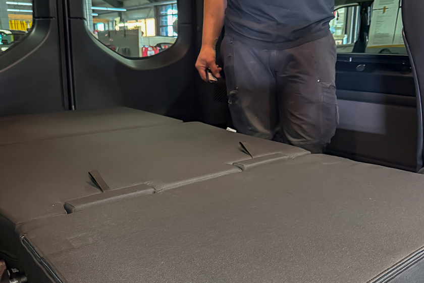 Eine Liegefläche in einem Auto, zum ausruhen und schlafen. Hinter der Liegefläche sieht man noch die Beine einer Person, die dort steht. 