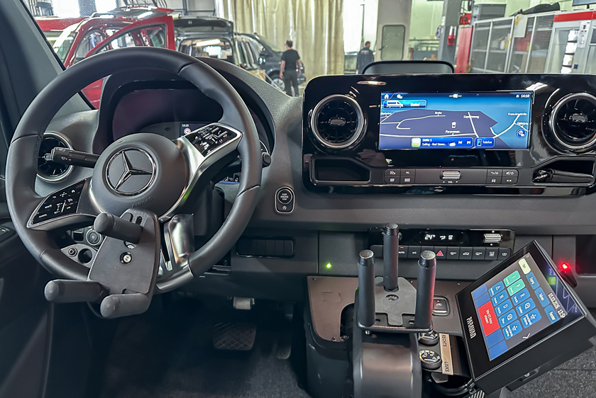 Links sieht man das Lenkrad eines Autos, welches mit einem Dreizack ausgestattet wurde. Rechts neben dem Lenkrad befindet sich ein weiterer Dreizack-Joystick, als Gas- Bremsschieber verbaut. Daran wurde das Paravan Touch Display gebaut. 