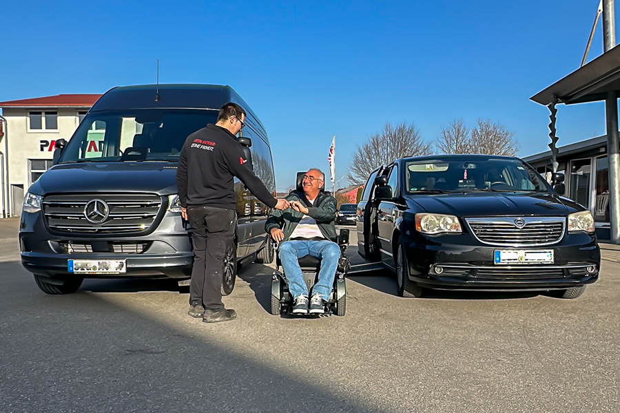 Bei blauem Himmel stehen zwei Fahrzeuge, links ein neuer Sprinter, rechts ein Chrysler Voyager auf dem Hof von Paravan. Zwischen den beiden Fahrzeugen sitzt ein Mann lachend im Rollstuhl, dem gerade ein Mitarbeiter von Paravan die Hand schüttelt.