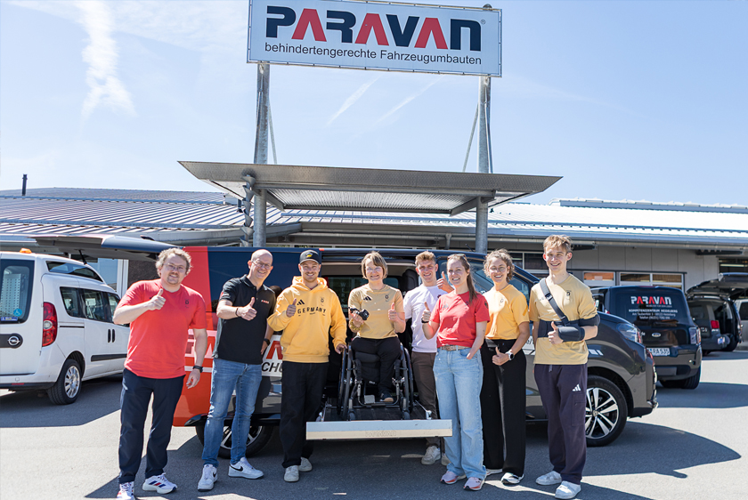 Mehrere Personen stehen vor einem Auto bei strahlendem Sonnenschein vor dem Paravan Mobilitätspark. Eine Dame im Rollstuhl steht auf dem herausgefahrenen Kassettenlift, weit oben damit sie gleich groß ist wie alle anderen. Bis auf zwei sind alle entweder in roten oder gelben Team Deutschland Klamotten auf dem Bild. 
