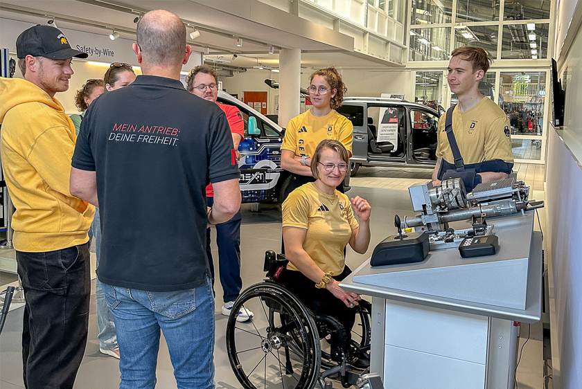 Mehrere Personen stehen in einem ungefährem Kreis, wobei ihnen ein Mitarbeiter der Paravan GmbH etwas erklärt. Rechts befindet sich ein Ausstellungstisch mit Joysticks...