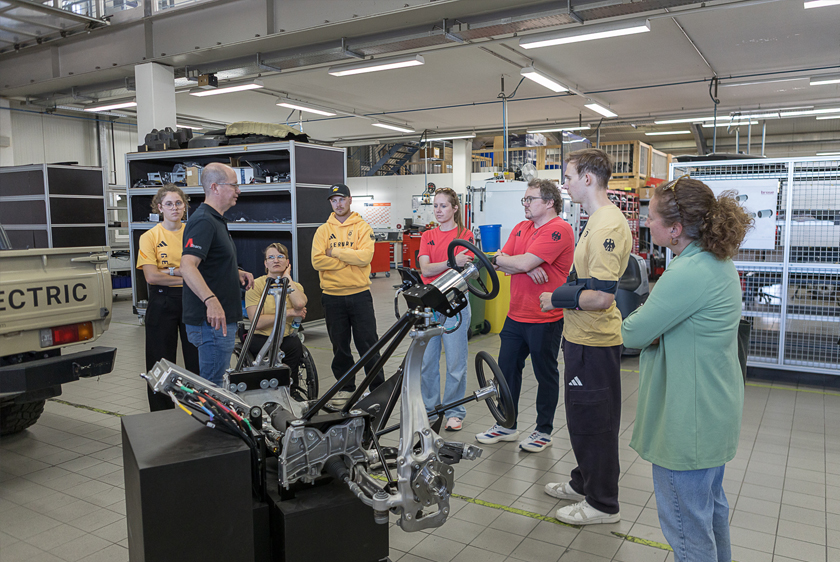 In einer Werkstatt steht eine Personengruppe aus ca. 8 Personen vor einem Ausstellungsstück, welches eine Fahrzeugachse mit Verbindung zum Lenkrad darstellt. Ein Mitarbeiter erklärt das ganze.
