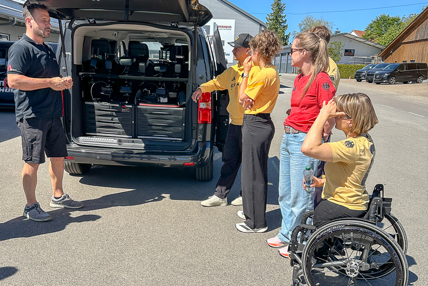 Links steht der Fahrlehrer der Paravan Fahrschule vor einem Fahrzeug mit geöffneter Heckklappe in dem sich verschiedene Joysticks befinden. Rechts vom Kofferraum stehen 6 Personen im Team Deutschland Outfit. Das ganze bei strahlendem Sonnenschein. 