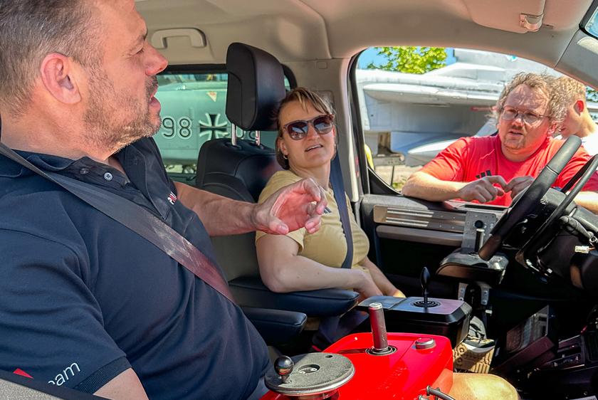 Zum geöffneten Fenster der Beifahrerseite sieht man zwei Personen im Fahrzeug sitzen sowie ein weiterer der zum geöffneten Fenster der Fahrerseite reinschaut. Auf der Beifahrerseite sitzt der Fahrlehrer der Paravan Fahrschule und auf dem Fahrersitz eine Dame mit Sonnenbrille. Alle drei Personen sind im Gespräch miteinander.