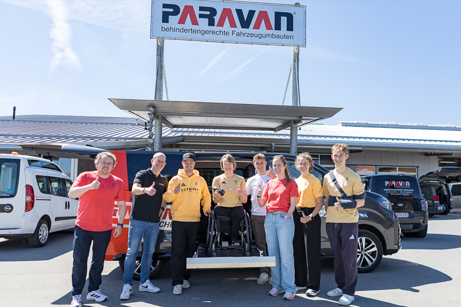 Mehrere Personen stehen vor einem Auto bei strahlendem Sonnenschein vor dem Paravan Mobilitätspark. Eine Dame im Rollstuhl steht auf dem herausgefahrenen Kassettenlift, weit oben damit sie gleich groß ist wie alle anderen. Bis auf zwei sind alle entweder in roten oder gelben Team Deutschland Klamotten auf dem Bild. 