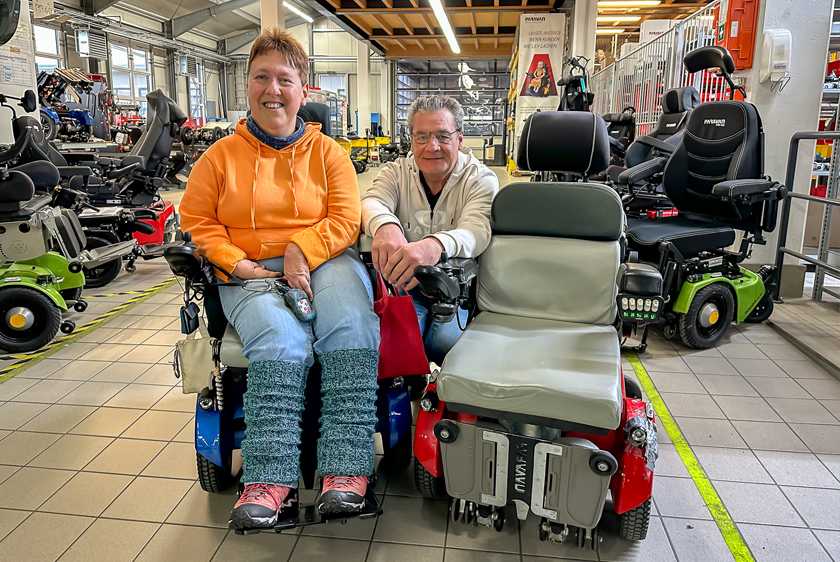 Eine Dame, mit orangenem Pullover, einem blauen neuen Rollstuhl, sowie rechts neben ihr, ein älterer roter Rollstuhl steht in der Rollstuhlwerkstatt der Paravan GmbH. In der Mitte hinter den beiden Rollstühlen kniet der Leiter der Rollstuhlabteilung. 