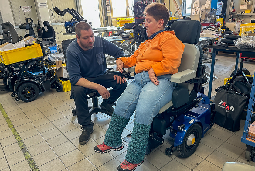 Eine Dame im orangenen Pullover sitzt in einem blauen PR50. Neben ihr sitzt ein Werkstatt Mitarbeiter der Rollstuhlabteilung von Paravan und schaut ihr zu. 