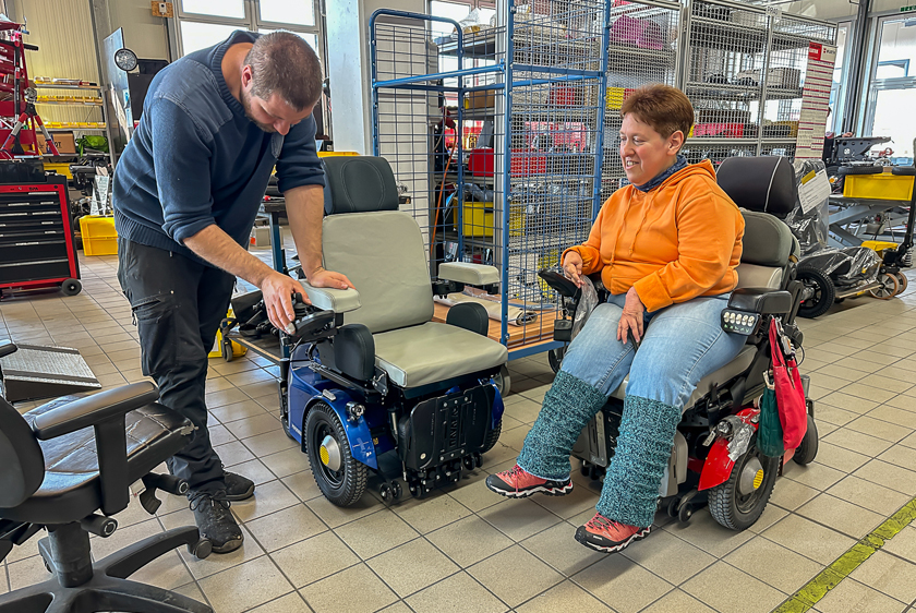 Ein Werkstatt Mitarbeiter der Paravan GmbH bearbeitet einen neuen blauen PR50 Elektrorollstuhl. Rechts daneben sitzt eine Dame mit orangenem Pullover ebenfalls in einem Rollstuhl und schaut dabei zu. 