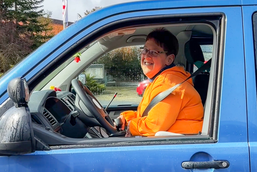 Lachend schaut eine Dame mit orangener Jacke aus dem Fahrerfenster eines blauen Autos heraus.