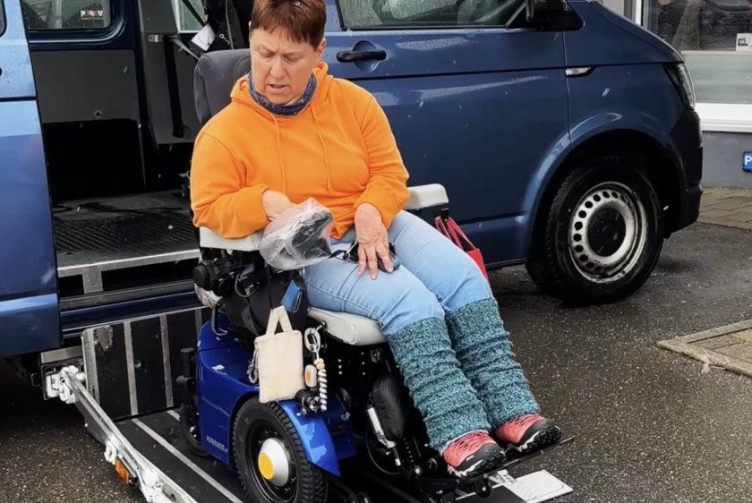 Eine Dame mit orangenem Pullover sitzt in ihrem blauen PR50 Rollstuhl. Der Rollstuhl steht auf einem herausgefahrenen Kassettenlift auf dem Boden vor einem blauen Auto. 