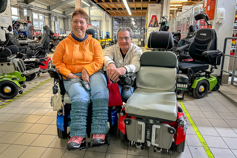 Eine Dame, mit orangenem Pullover, einem blauen neuen Rollstuhl, sowie rechts neben ihr, ein älterer roter Rollstuhl steht in der Rollstuhlwerkstatt der Paravan GmbH. In der Mitte hinter den beiden Rollstühlen kniet der Leiter der Rollstuhlabteilung. 