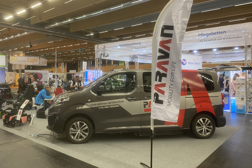 Ein Messestand der Paravan GmbH auf dem sich mittig im Vordergrund eine Paravan Beachflag befindet. Dahinter ein Auto, sowie Links neben dem Auto ein paar Rollstühle. 