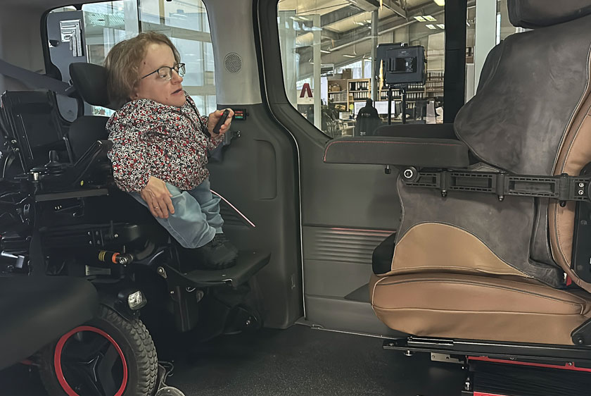 Eine Dame mit Kleinwuchs befindet sich im Auto mit ihrem Rollstuhl, gegenüber befindet sich der Fahrersitz, jedoch umgedreht, damit sie aus dem Rollstuhl auf den Fahrersitz Umsitzen kann.