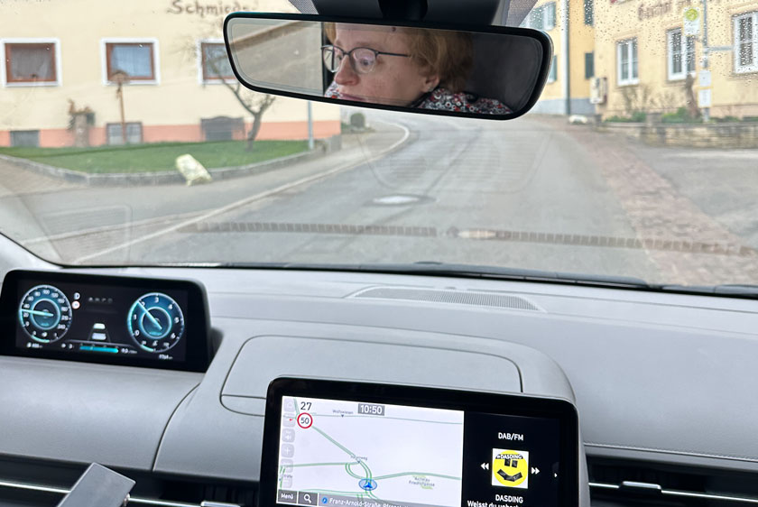 Ein Blick aus dem Auto zur Frontscheibe hinaus auf die Straße in einer Ortschaft. Durch die Spiegelung im Rückspiegel sieht man eine konzentrierte Dame.