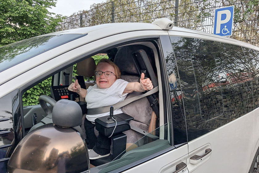 Eine lachende Dame streckt ihren rechten Arm zum geöffneten Fahrerfenster mit Daumen hoch hinaus. In ihrem Fahrzeug befinden sich zwei Joysticks verbaut, ein Paravan Touch Display und ein individuell angepasster Fahrersitz. Rechts oben am Bildrand sieht man ein Parkplatzschild für Behinderte Personen.
