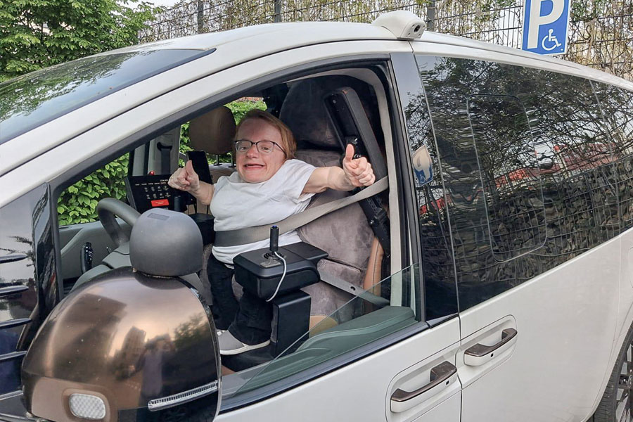 Eine lachende Dame streckt ihren rechten Arm zum geöffneten Fahrerfenster mit Daumen hoch hinaus. In ihrem Fahrzeug befinden sich zwei Joysticks verbaut, ein Paravan Touch Display und ein individuell angepasster Fahrersitz. Rechts oben am Bildrand sieht man ein Parkplatzschild für Behinderte Personen.