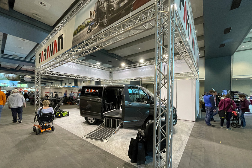 Ein Messestand mit Traversensystem, auf dem ein Auto mit Paravan Logo, sowie Rollstühle stehen.