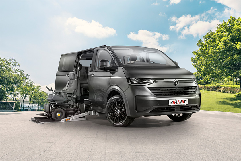 Eine VW Caravelle steht in einer animierten Umgebung mit einem großen Platz und rechts davon einem Park. Ein schwarzer Rollstuhl steht auf dem herausgefahrenen Kassettenlift, bei geöffneter Seitentüre de VW Caravelle. 