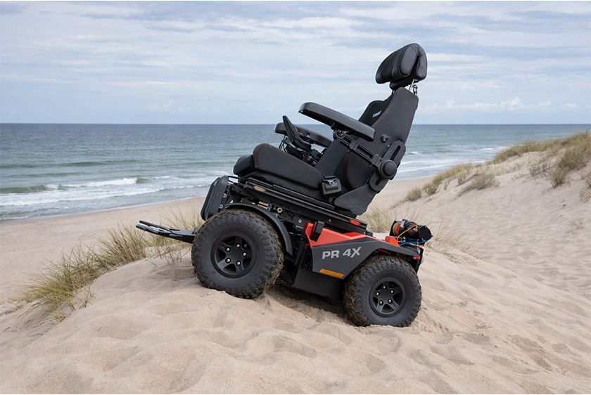 Der PR4X Offroad Elektrorollstuhl, steht mitten im Sand auf einer Düne. Links befindet sich das Meer.