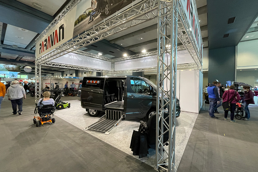 Ein Messestand mit Traversensystem, auf dem ein Auto mit Paravan Logo, sowie Rollstühle stehen.