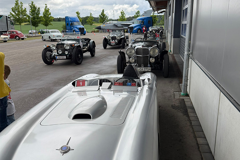 Mehrere Oldtimer Autos stehen nebeneinander, sowie hintereinander links neben einer Halle. Im Hintergrund stehen zwei blaue Trucks, sowie ein Tornado.