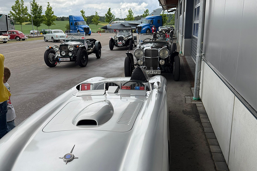 Mehrere Oldtimer Autos stehen nebeneinander, sowie hintereinander links neben einer Halle. Im Hintergrund stehen zwei blaue Trucks, sowie ein Tornado.