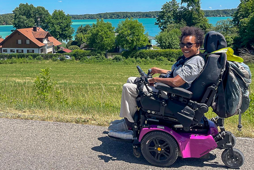 Eine ältere strahlend lachende Dame im pinken PR30/II Elektrorollstuhl, unterwegs am Tegernsee bei strahlendem Sonnenschein und grünen Wiesen.